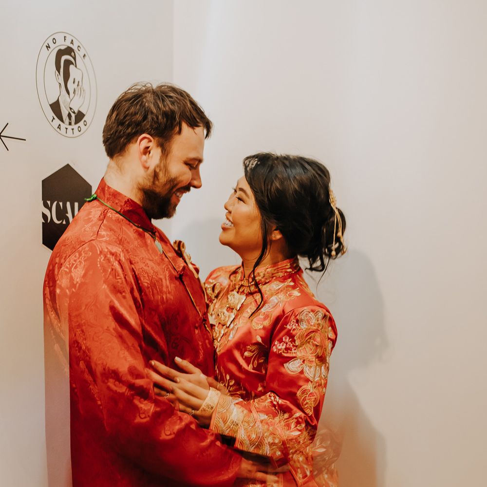 Bride in red and gold traditional Chinese tea ceremony dress stands with groom in black trousers and red traditional Chinese clothing