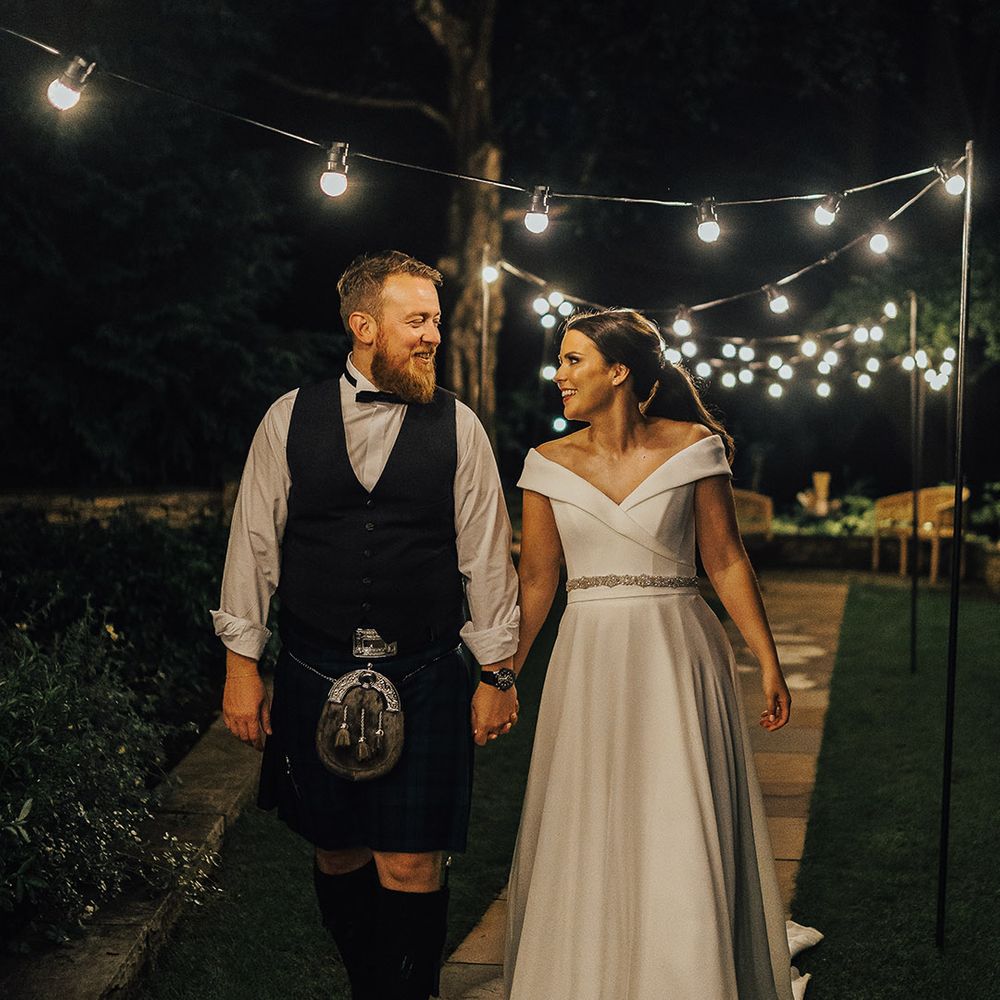Bride in an off the shoulder wedding dress and groom in a tartan kilt holding hands under a canopy of festoon lights at Foxhill Manor 
