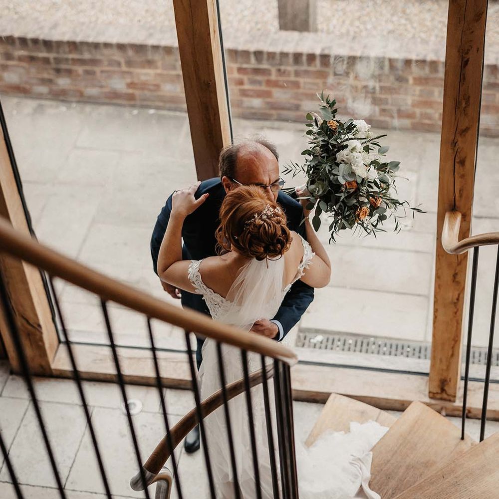 Bride holding white and orange wedding bouquet walks down the stairs to meet her father who wears blue suit