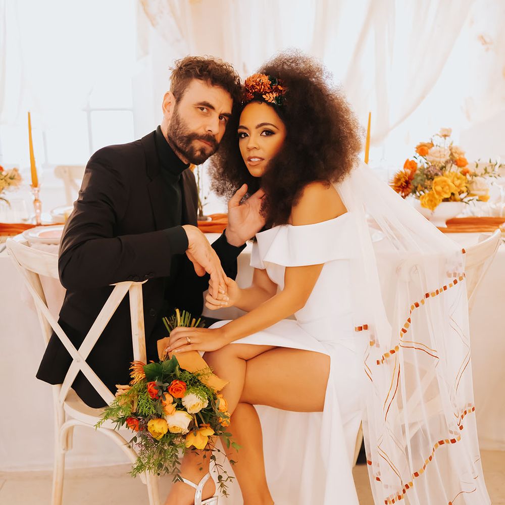 Black bride in an off the shoulder wedding dress with front slit and patterned veil sitting at her reception table with her groom in a black suit holding an orange and yellow golden wedding bouquet 