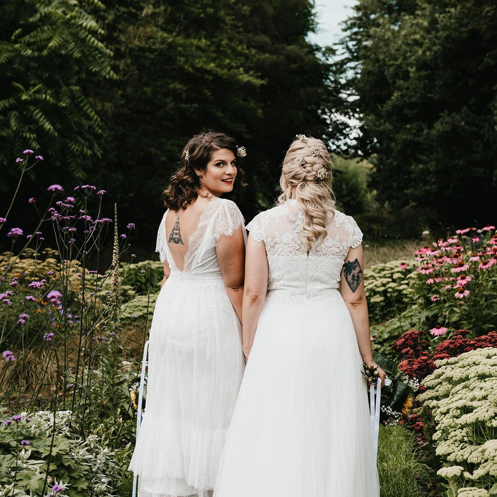 Bride with brunette hair looks back at the camera as she walks with bride with blonde hair through the gardens of Chaucer Barn venue
