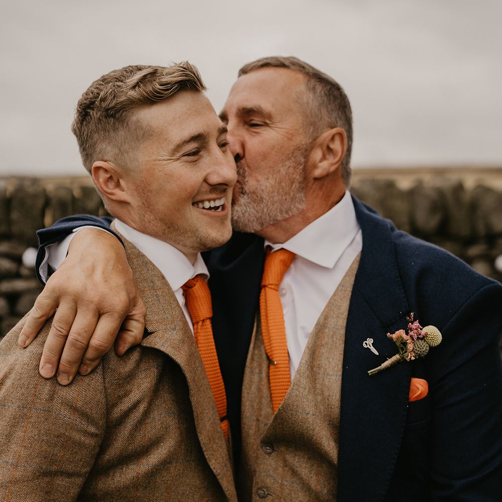 Father in dark suit jacket and brown waistcoat kisses groom on the cheek in matching brown suit and orange tie