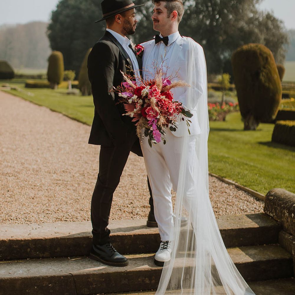 Groom in a black suit and fedora hat standing with his partner at Prestwold Hall Barns in a white groom suit and cape holding a vibrant red bouquet 