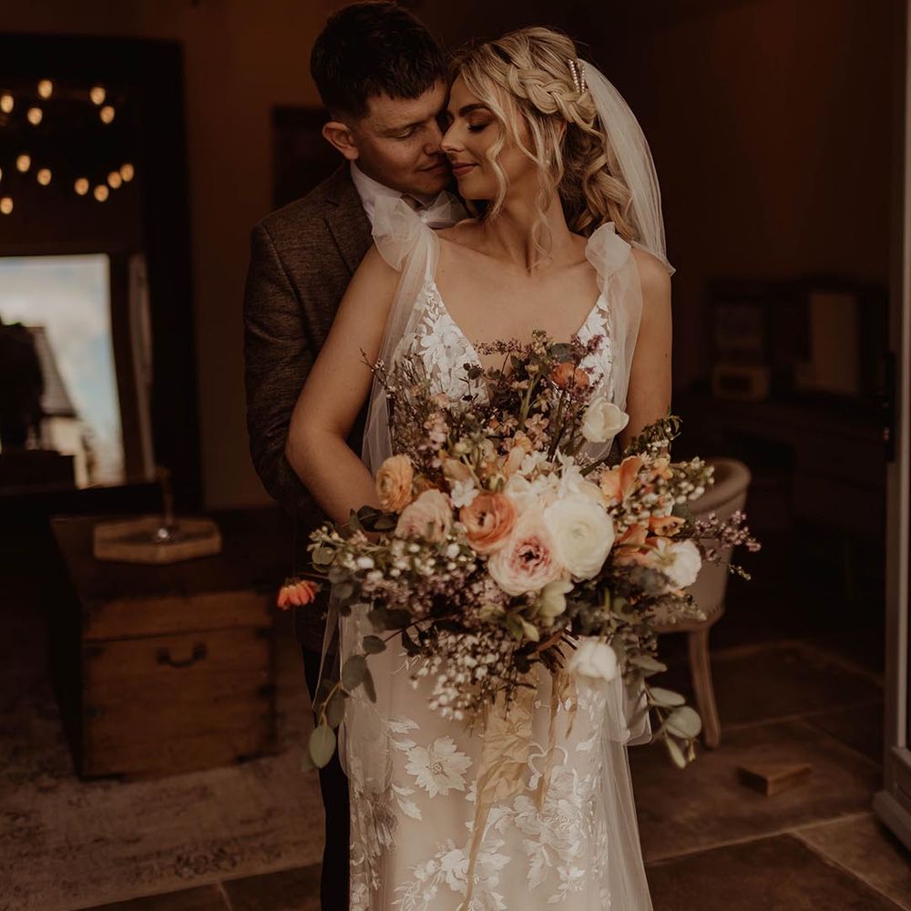 Bride in Made With Love lace wedding dress with bow sleeves holding autumnal wedding bouquet being hugged from behind by the groom in brown suit for rustic wedding 