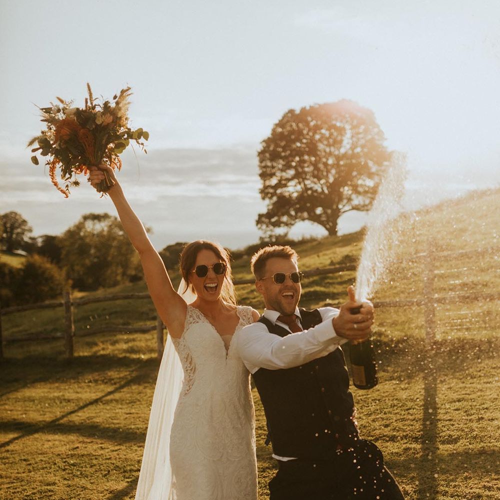 Bride in lace dress with long veil and neutral bouquet with groom doing champagne pop