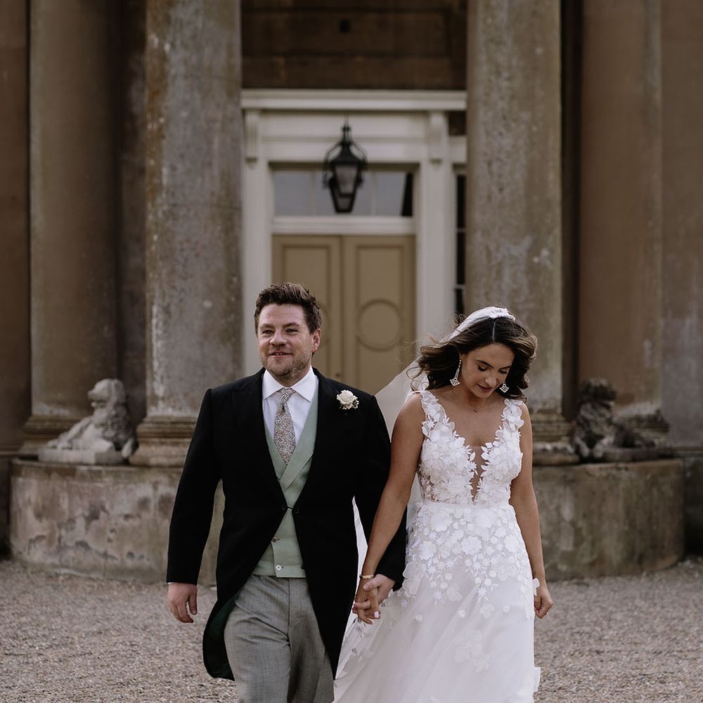 Bride in floral applique wedding dress walks around holding hands with the groom in morning suit with green waistcoat 