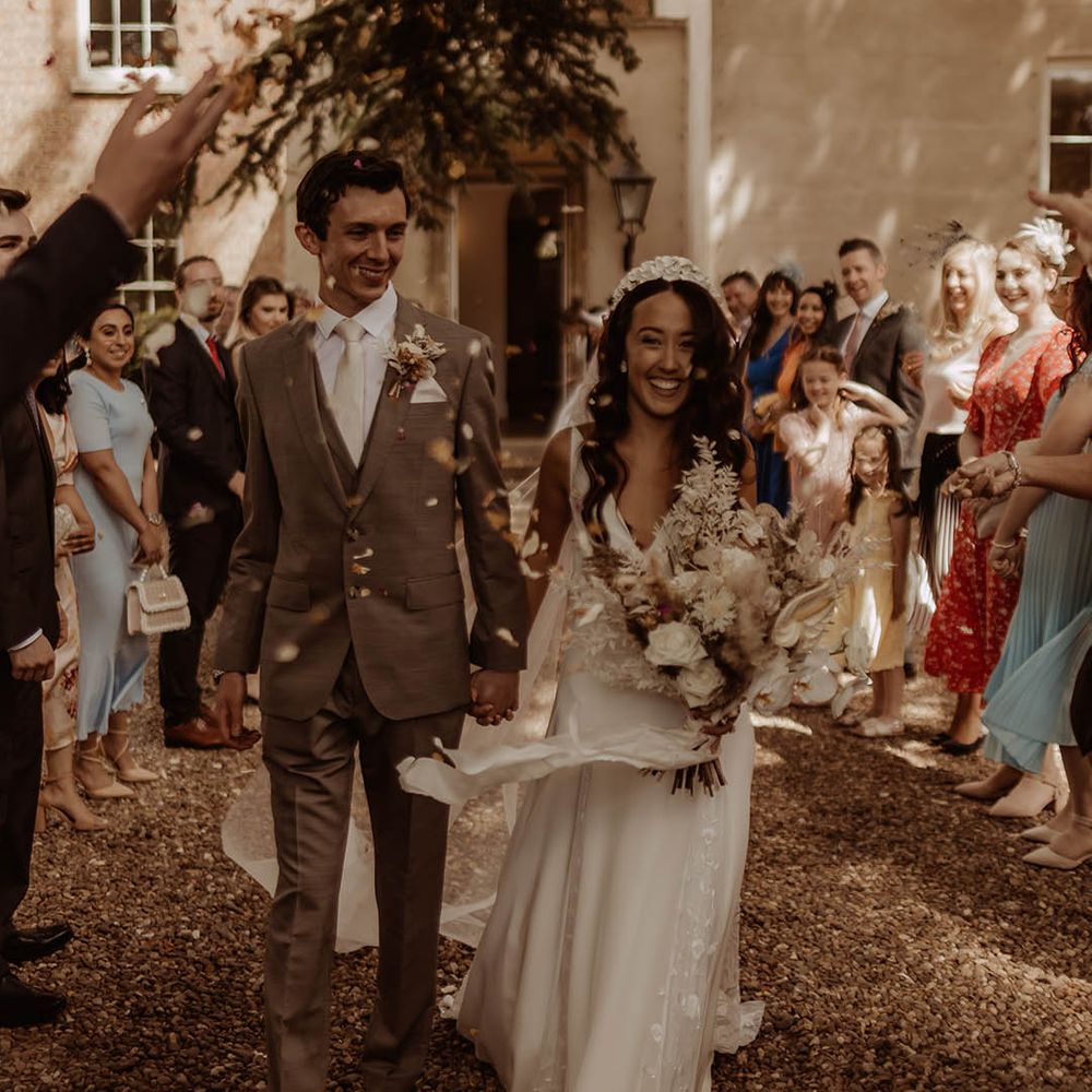 Bride wearing a bridal headband carrying a white flower dried grass bouquet and groom in grey suit have confetti exit from wedding 