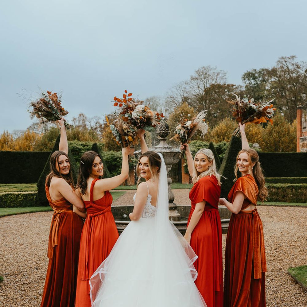 Bridesmaids in burnt orange dresses with bride in layered tulle princess skirt all holding matching autumnal bouquets 