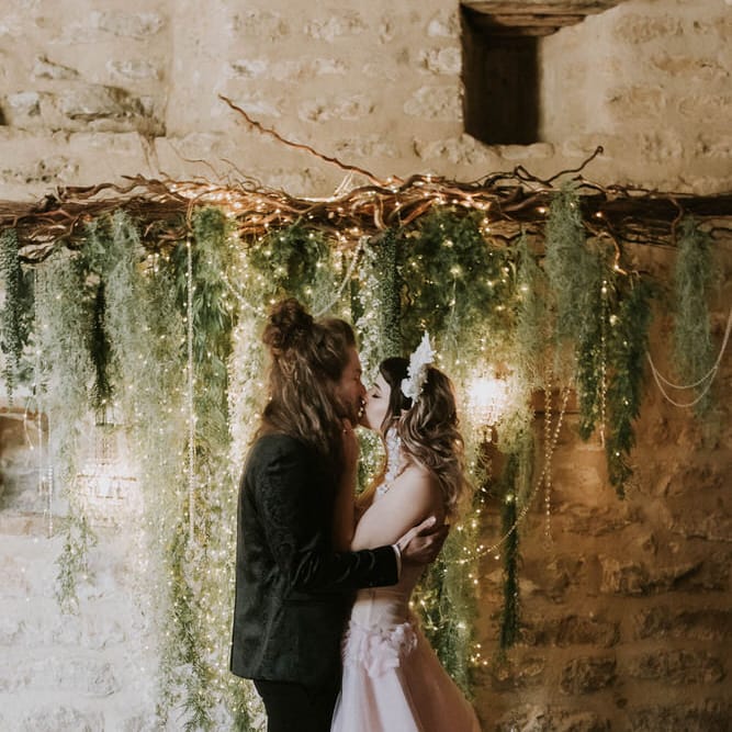 Groom in a tuxedo and bride in a pink strapless wedding dress kissing at the altar with a twig, fairy light and foliage backdrop 
