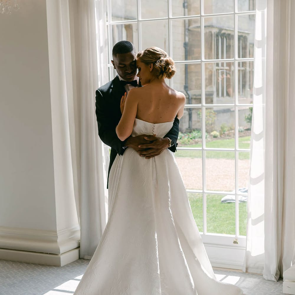 Bride in strapless and pleated brocade Jesus Peiro wedding dress hugging the groom as they stand next to a window 