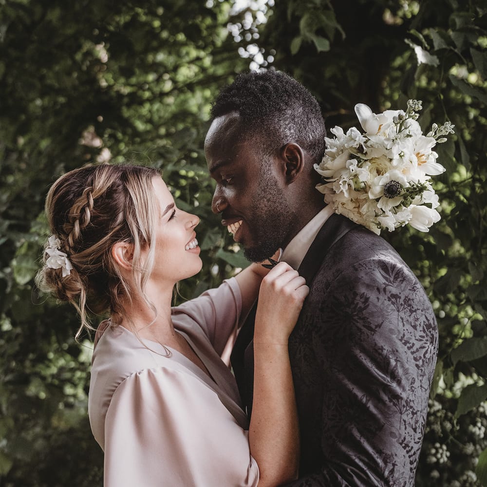 Bridesmaid in pale pink dress hugs a groomsmen in a black patterned jacket with a white wedding bouquet