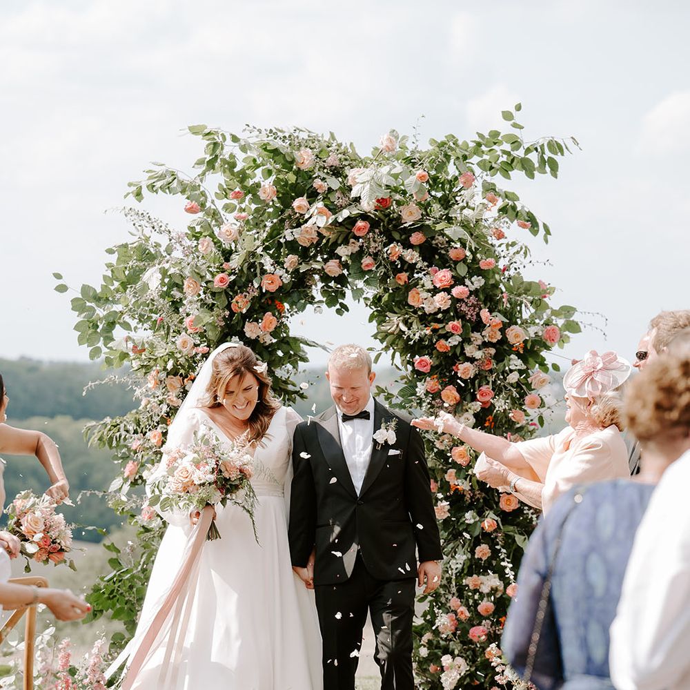 Groom in black tie walks through white petals with his groom after outdoor wedding ceremony with floral arch