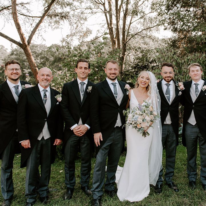 Bride in Made With Love wedding dress and flower applique veil with the groom, groomsmen and family members in three piece suits with grey waistcoats and blue ties with clips
