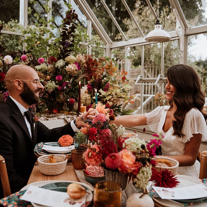 Bride and groom holding hands over neutral, botanical and autumnal wedding tablescape with neutral toned tablerunner on rustic wooden table, orange, green and red coloured glasses, earthenware, seasonal fruit and vegetables, red, brown and green tapered candles in gold candlesticks and clear vases with autumnal coloured flowers