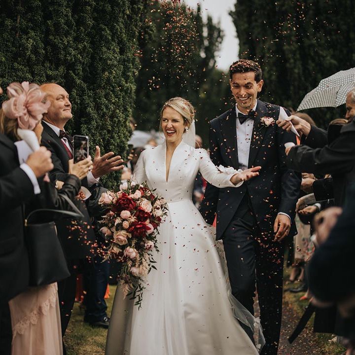 Bride holding pink and red cascading bouquet walking with the groom in black tie as the guests throw confetti over the couple