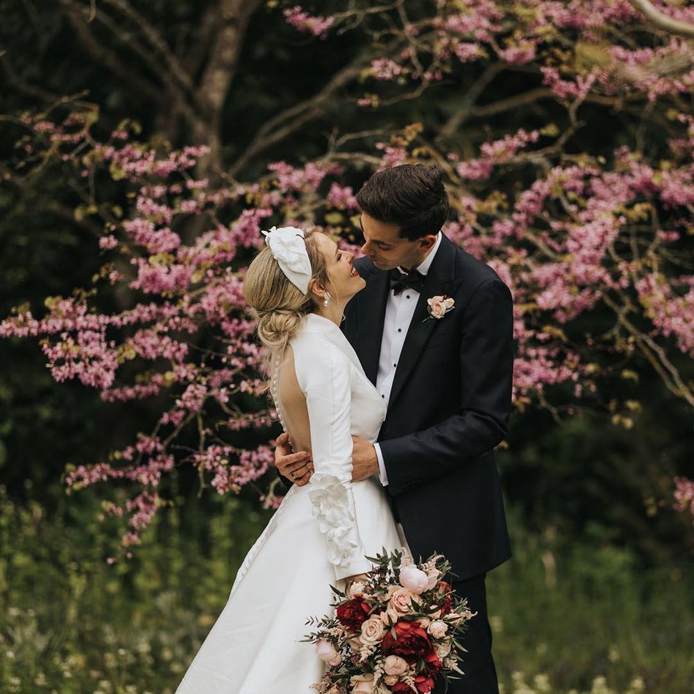 Bride in button back illusion wedding dress with 3d flower detailing on the long sleeves with a collar and flower hairpiece leans in for a kiss with the groom in black tie 