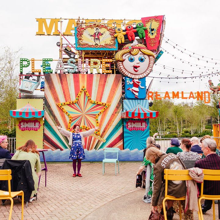 Wedding celebrant wearing blue patterned dress and cover up giving speech at theme park wedding with rides, rollercoasters and Ferris wheels in the back 