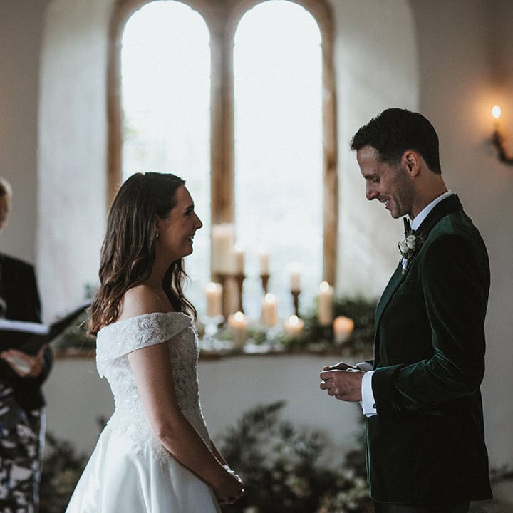 Groom in a dark green velvet tuxedo smiles as he reads out his wedding vows to the bride standing opposite him in an off the shoulder lace wedding dress