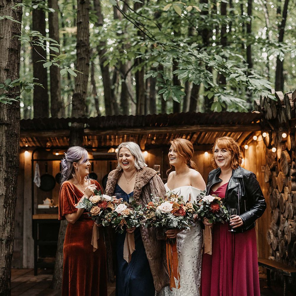 Bride stands beside her bridesmaids in mismatched Autumnal bridesmaid dresses with dried floral bouquets 