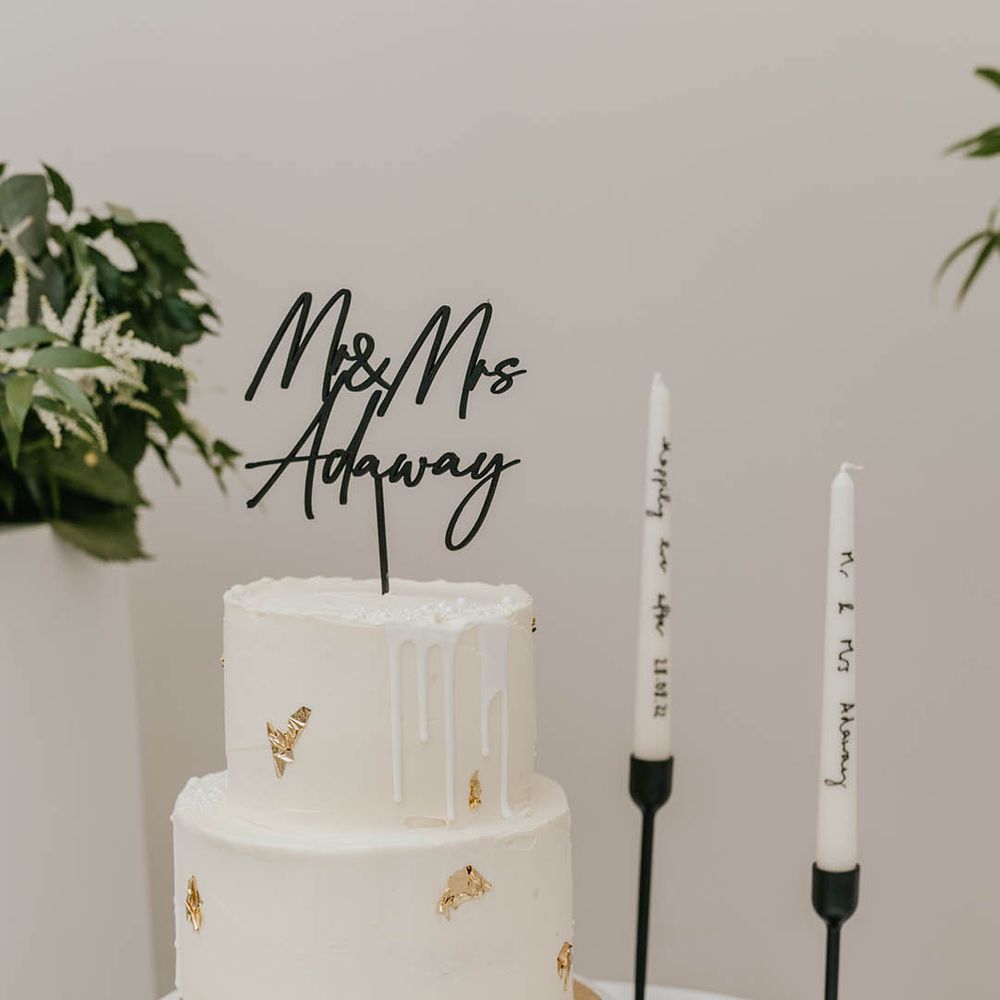 Two tier white frosted wedding cake with drip icing, pearls, and gold flakes with black metal personalised cake topper with black candle holders and white personalised taper candles