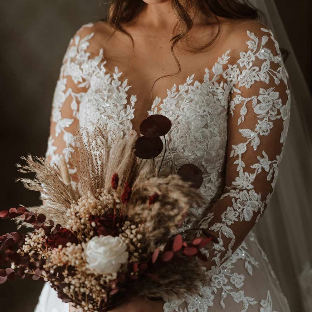 Bride wearing long sleeve lace illusion sleeve wedding dress with overlay holding large autumnal boho dried flower bridal bouquet 