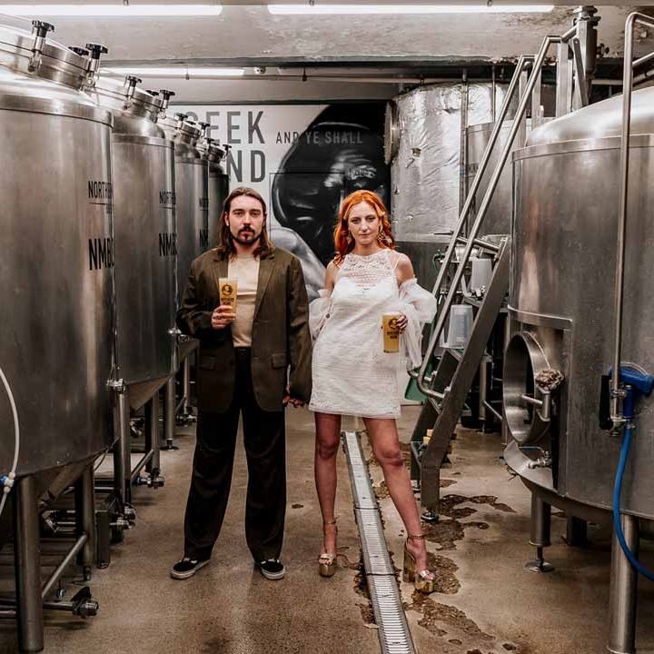 Groom in brown blazer, white shirt, grey suit trousers and vans shoes standing with bride in 70s inspired boatneck short lace wedding dress with detachable tulle puff sleeves and platform gold wedding heels holding pints of beer by beer fermenters