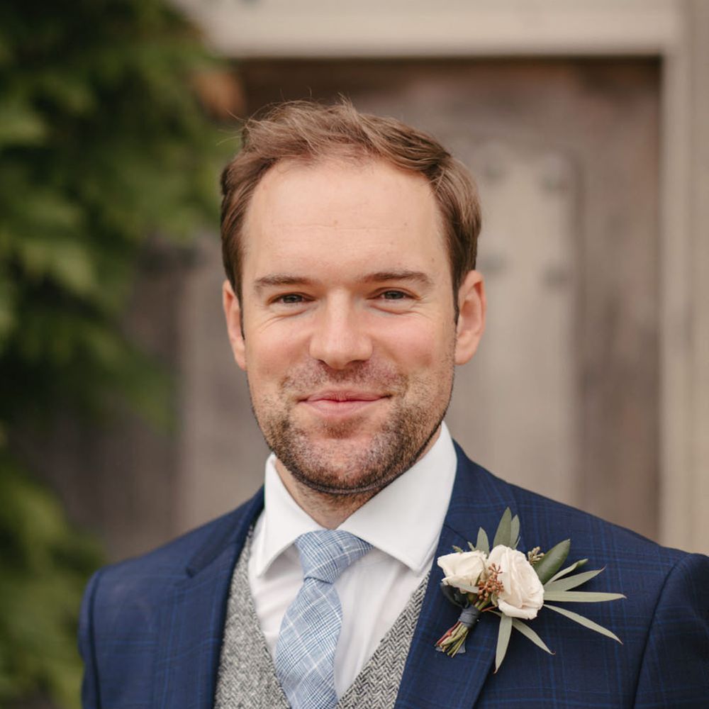 Groom wearing a navy blue wedding suit with a light grey waistcoat and blue tie with floral buttonhole accessory 