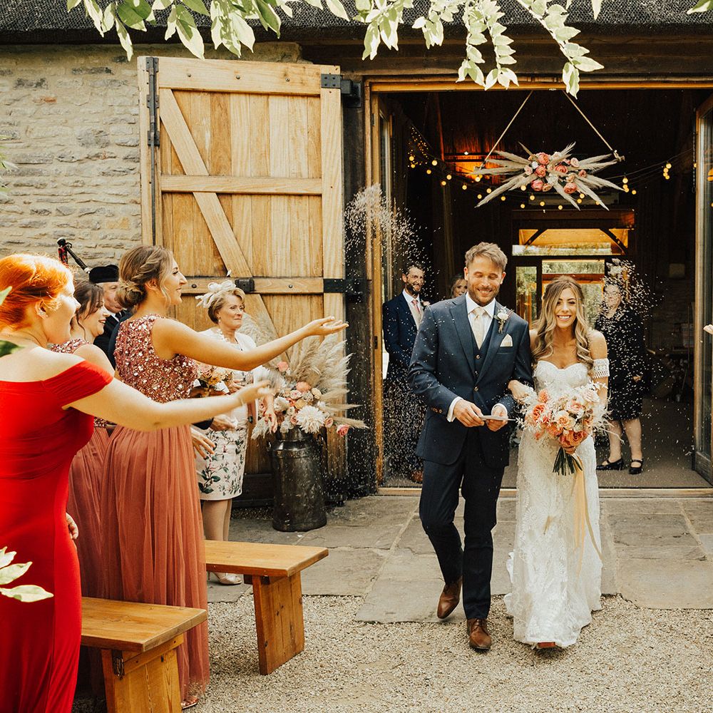 Boho bride holding rose bouquet and groom in navy suit walk under confetti at Tythe Barn wedding