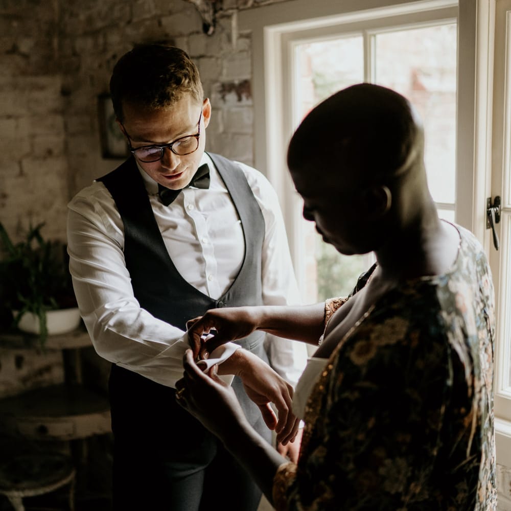 Black bride with alopecia helping her groom put on his cufflinks on the wedding morning 