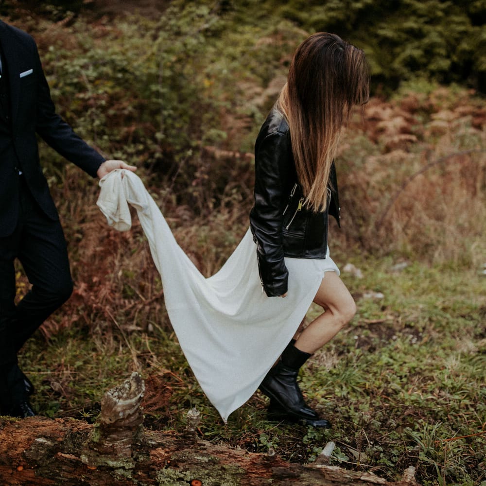 The groom holding the brides skirt off the ground as they walk through the woods