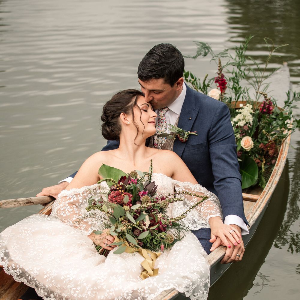 Bride & groom sit in boat as it floats along the river whilst bride holds bouquet