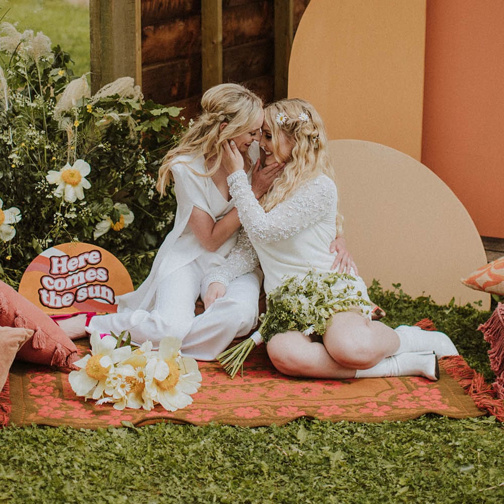 Two brides sitting on a wool rug with retro 'Here Comes The Sun' wedding sign and colour block altar decor 
