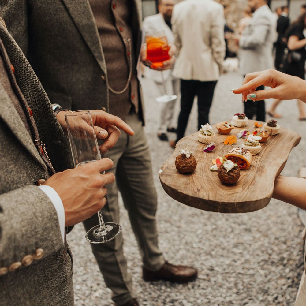 Waitress holds wooden board with assorted canapés on as guests in sage green suits holding drinks have a look in courtyard during wedding reception at Anran Devon