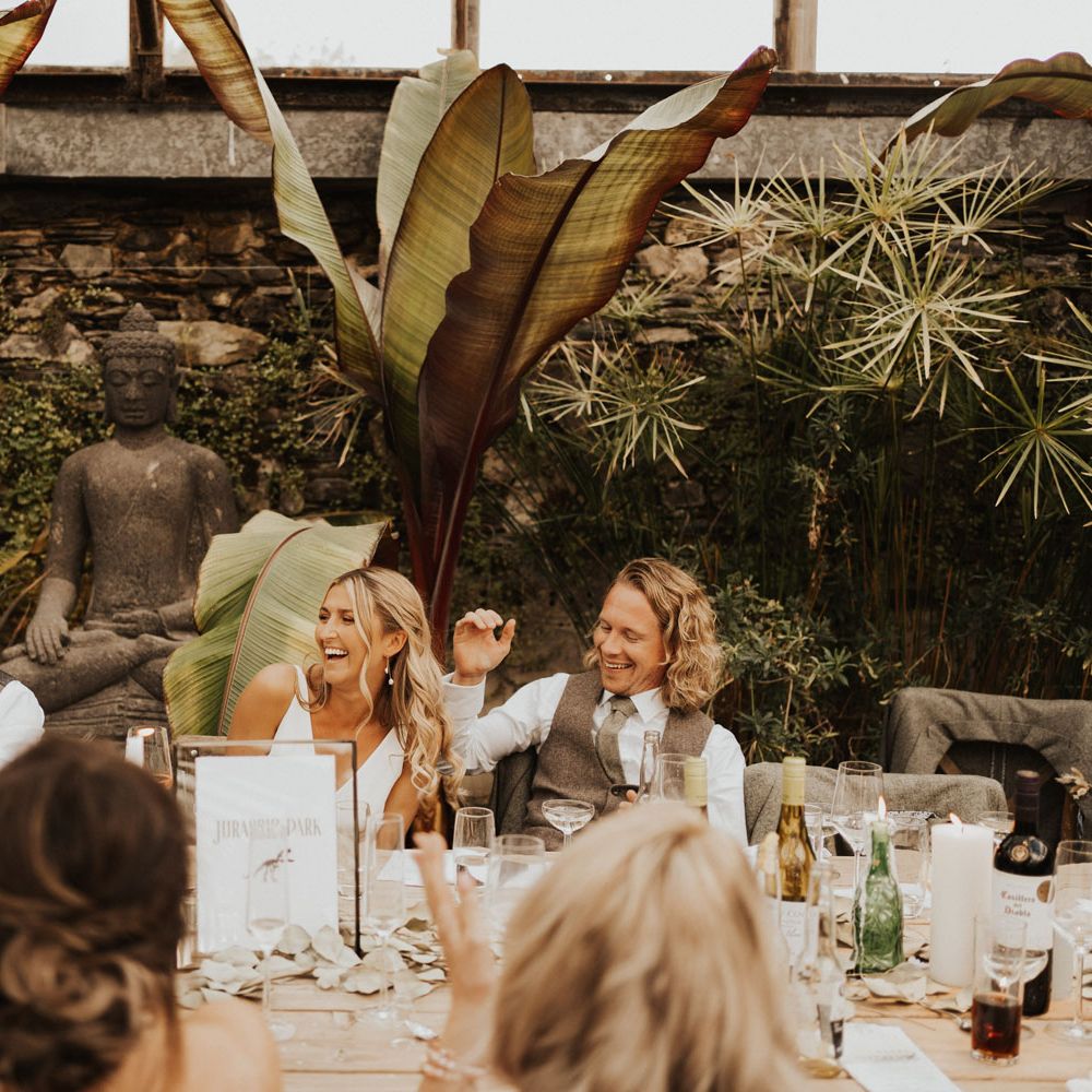 Bride in white Made With Love wedding dress laughs with groom in sage green waistcoat and tie and guests as they listen to speeches whilst sat at wooden table in the glasshouse at Anran Devon during wedding breakfast