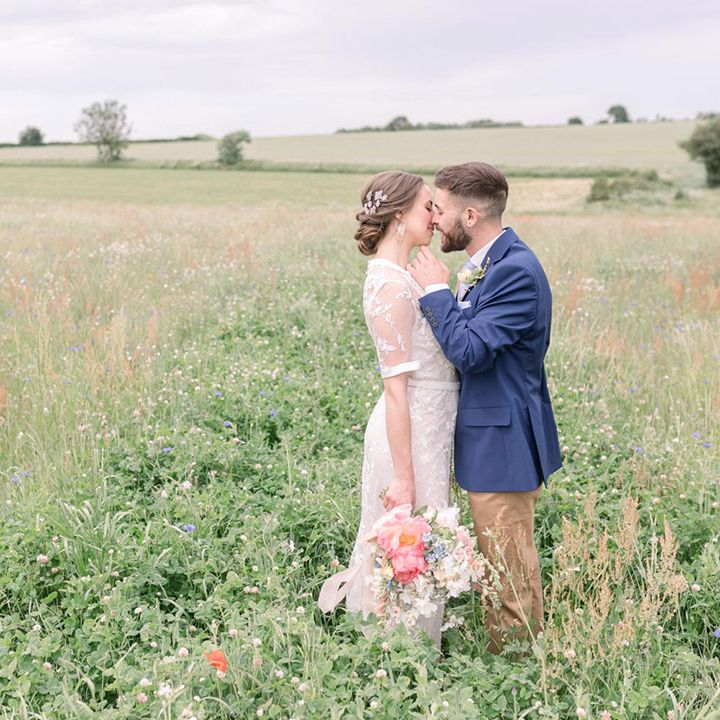 Groom in beige chinos and a navy jacket kissing his bride in a meadow as she wears a lace wedding dress and carries a pastel flower bouquet 