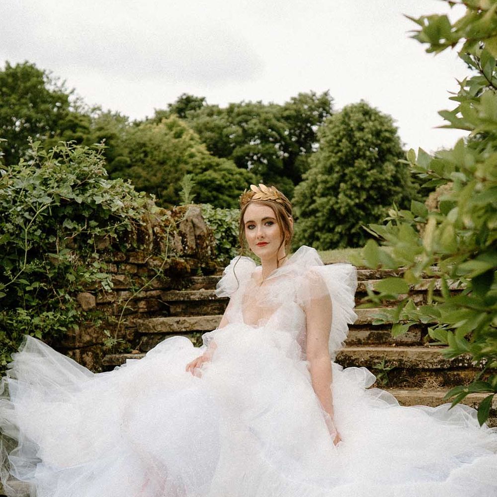 Beautiful bride in a tulle wedding dress with embellished wedding shoes wearing a gold crown sitting on the steps at Bourton Hall 
