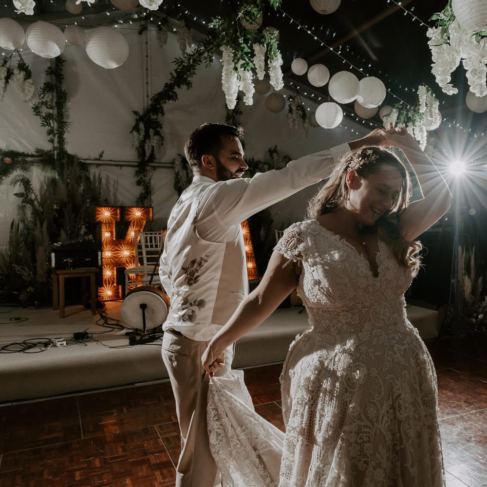 Bride in lace homemade wedding dress with train is twirled around dance floor by groom in beige waistcoat and suit trousers inside wedding reception marquee with garlands, paper lanterns and fairy lights at garden wedding in Bedfordshire