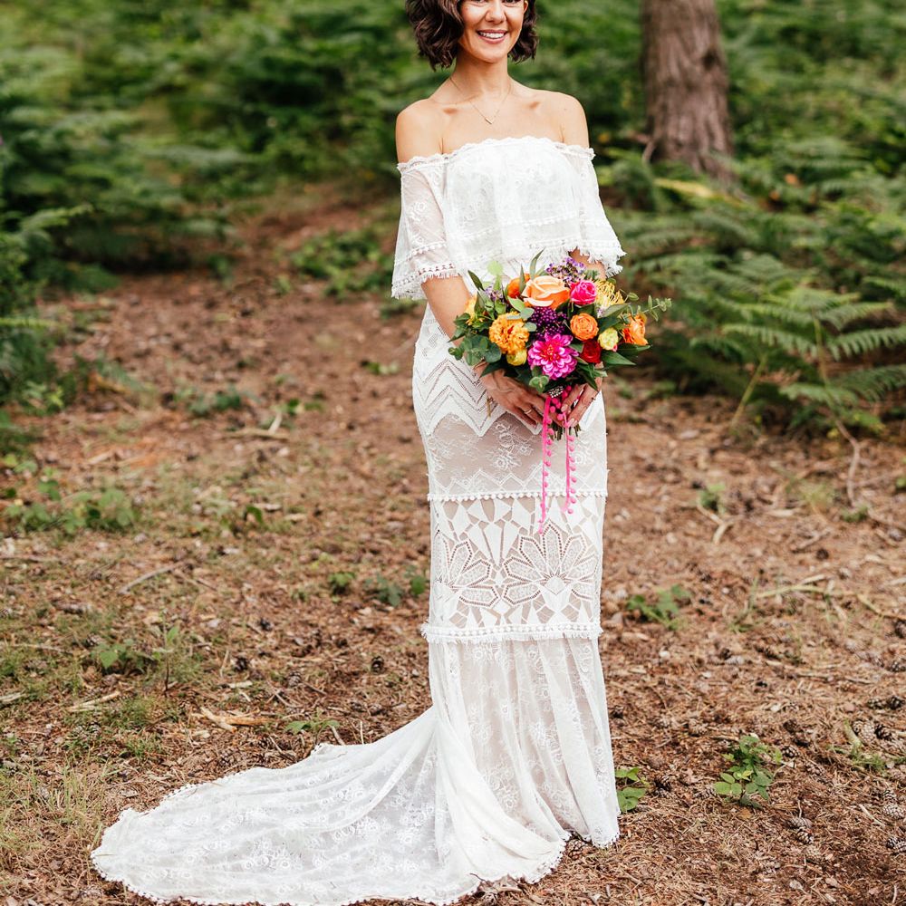 Bride in off the shoulder Grace Loves Lace wedding dress and pink and orange flower crown stands in woodland holding colourful bridal bouquet after woodland wedding ceremony at The Bridal Barn