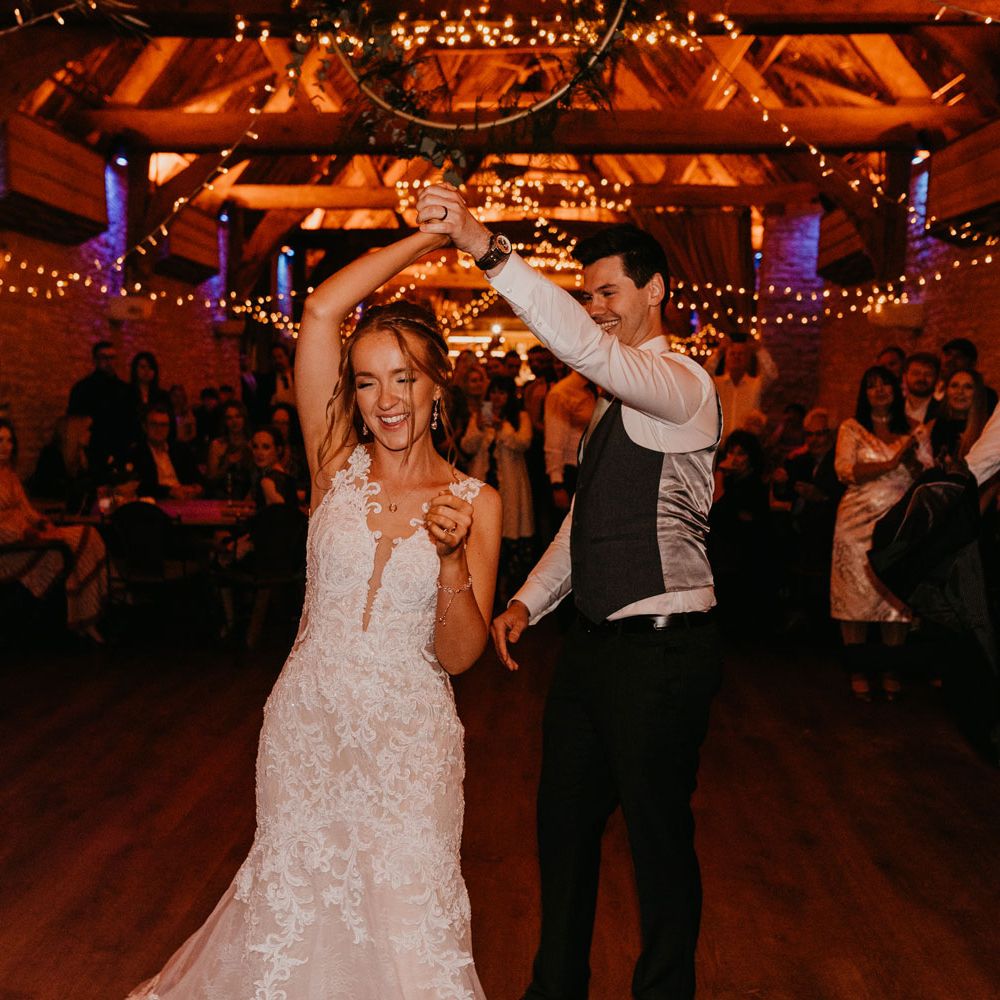 Groom in white shirt and dark waistcoat twirls bride in lace wedding dress around on the dance floor during rustic barn wedding reception lit by fairy lights