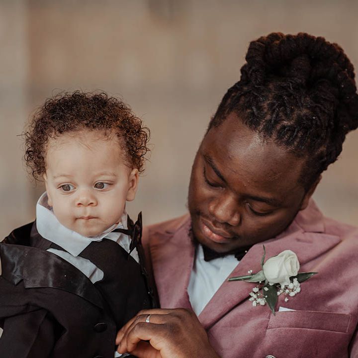 Groom holds his son on his wedding day who wears suit with black tie 