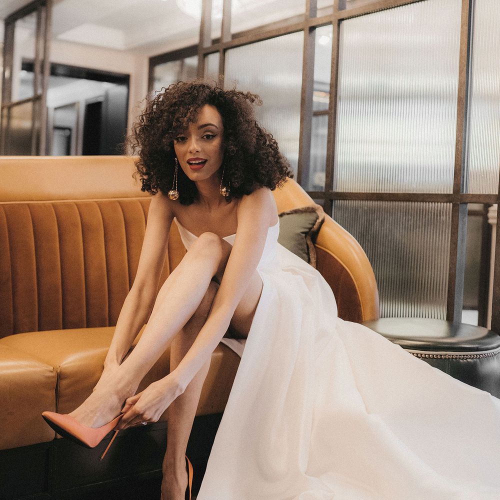 Black bride in a. strapless wedding dress with naturally curly hair, red lipstick and dangly earrings putting on her orange stiletto wedding shoes 