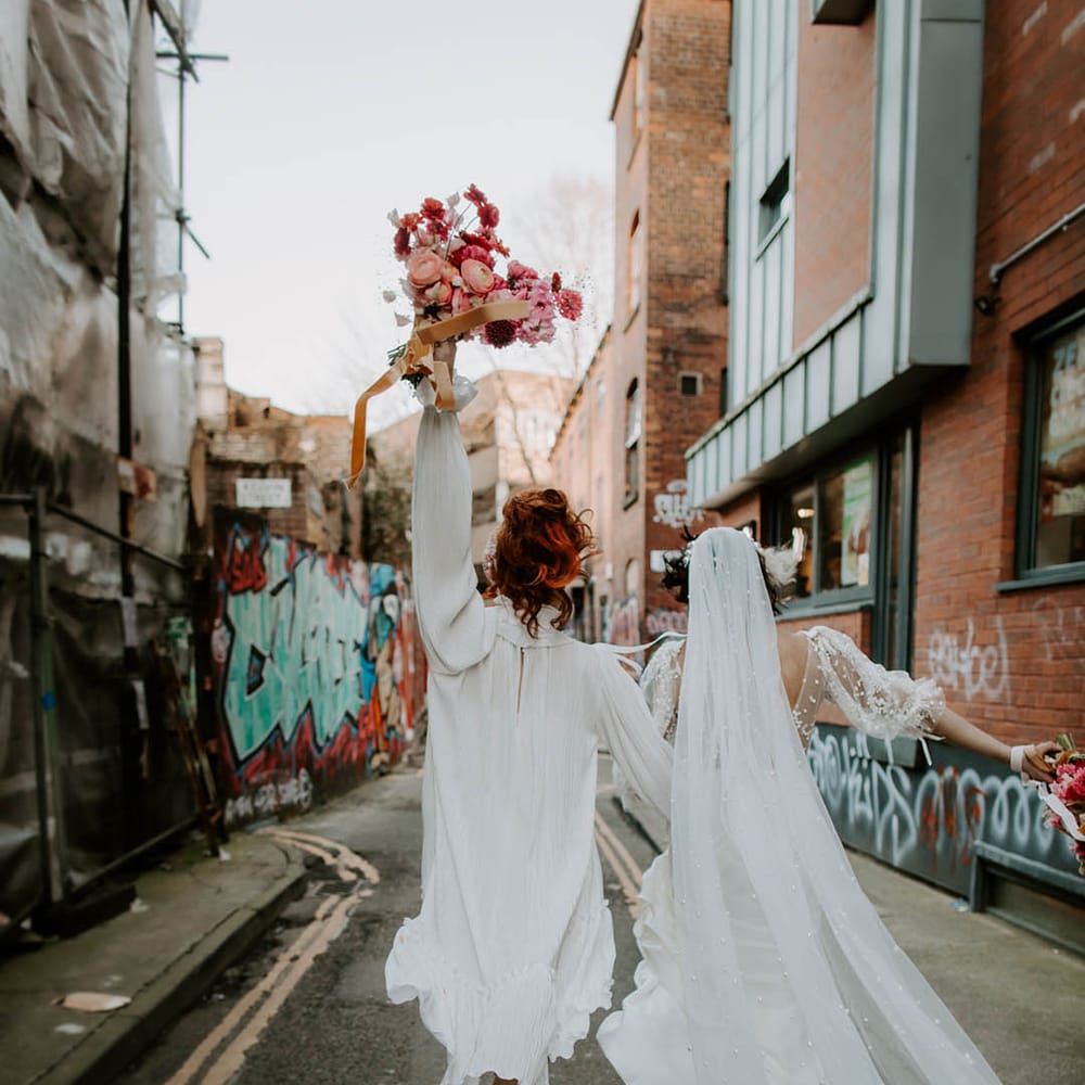 Bride in a short wedding dress holding her red and pink bouquet in the air arm in arm with her bride in a long pearl embellished wedding veil 