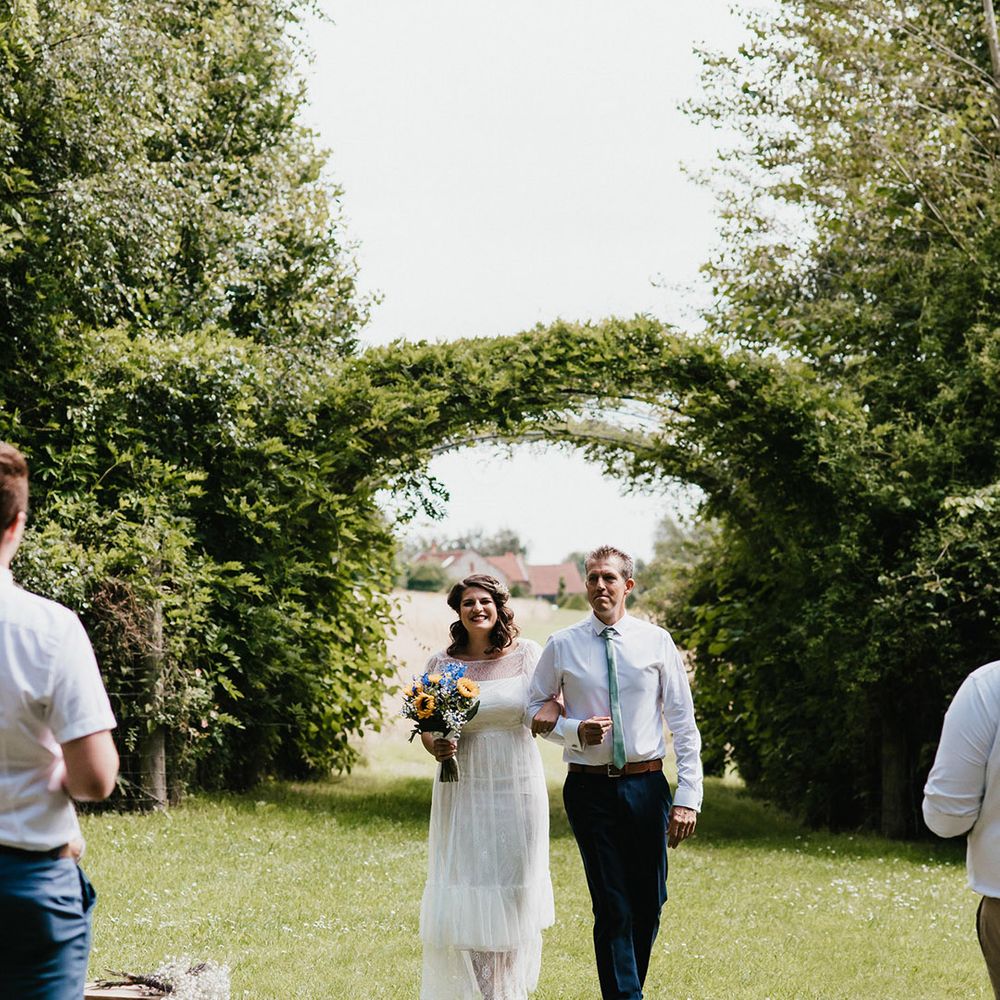 Bride walked down the aisle by the father of the bride in white shirt and green tie with dark trousers for outdoor ceremony 