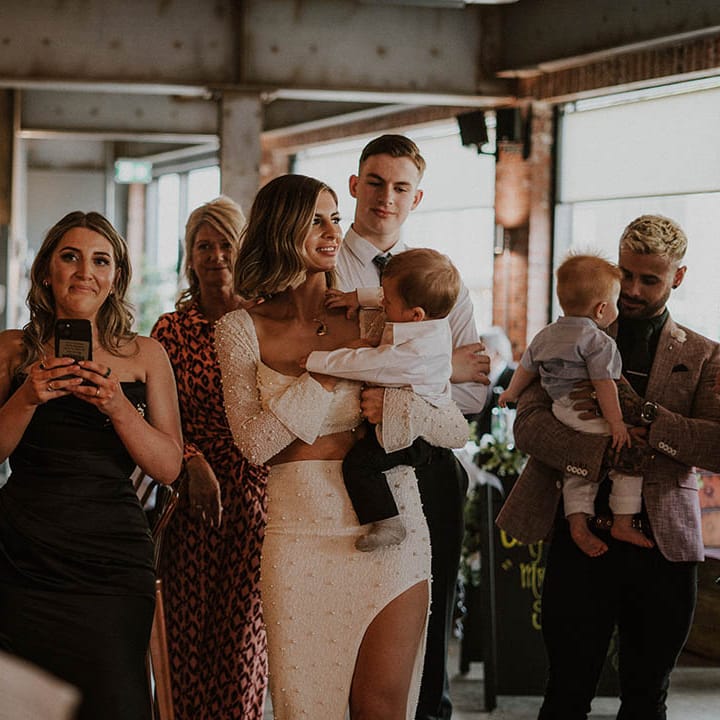 Bride in white pearl co ord holding son with wedding guests 