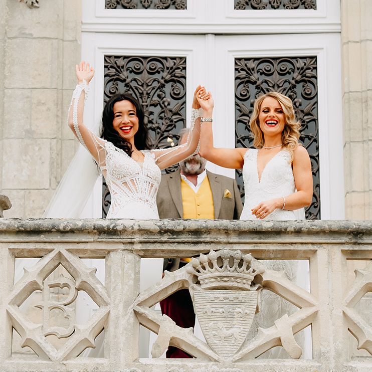 Brides raise their joint hands for guests to see as they are married with both wearing matching red lipstick and lace dresses 