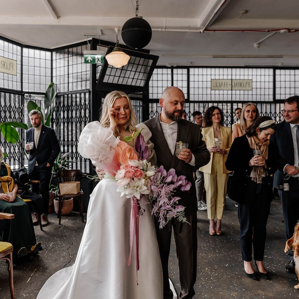 Bride in a JESUS PEIRO wedding dress holding a pastel wedding bouquet crying with her husband in a doubled breasted jacket during the drinks reception speeches 