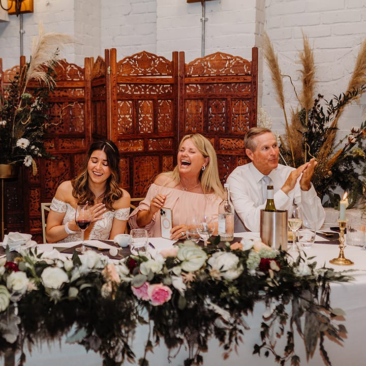 Bride sits with wedding guests at the wedding reception with a boho and rustic style with the wooden screen and dried flowers 
