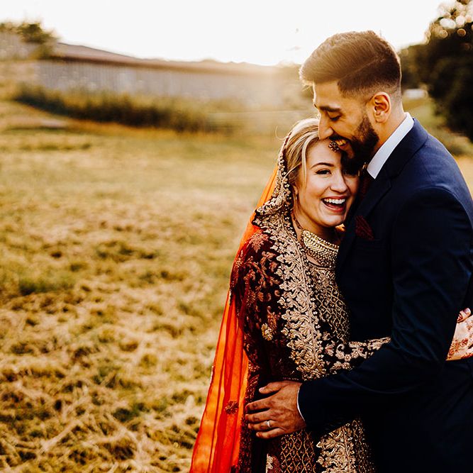 Bride wearing brightly coloured saree with gold embellishment embraces her groom outdoors in field during golden hour