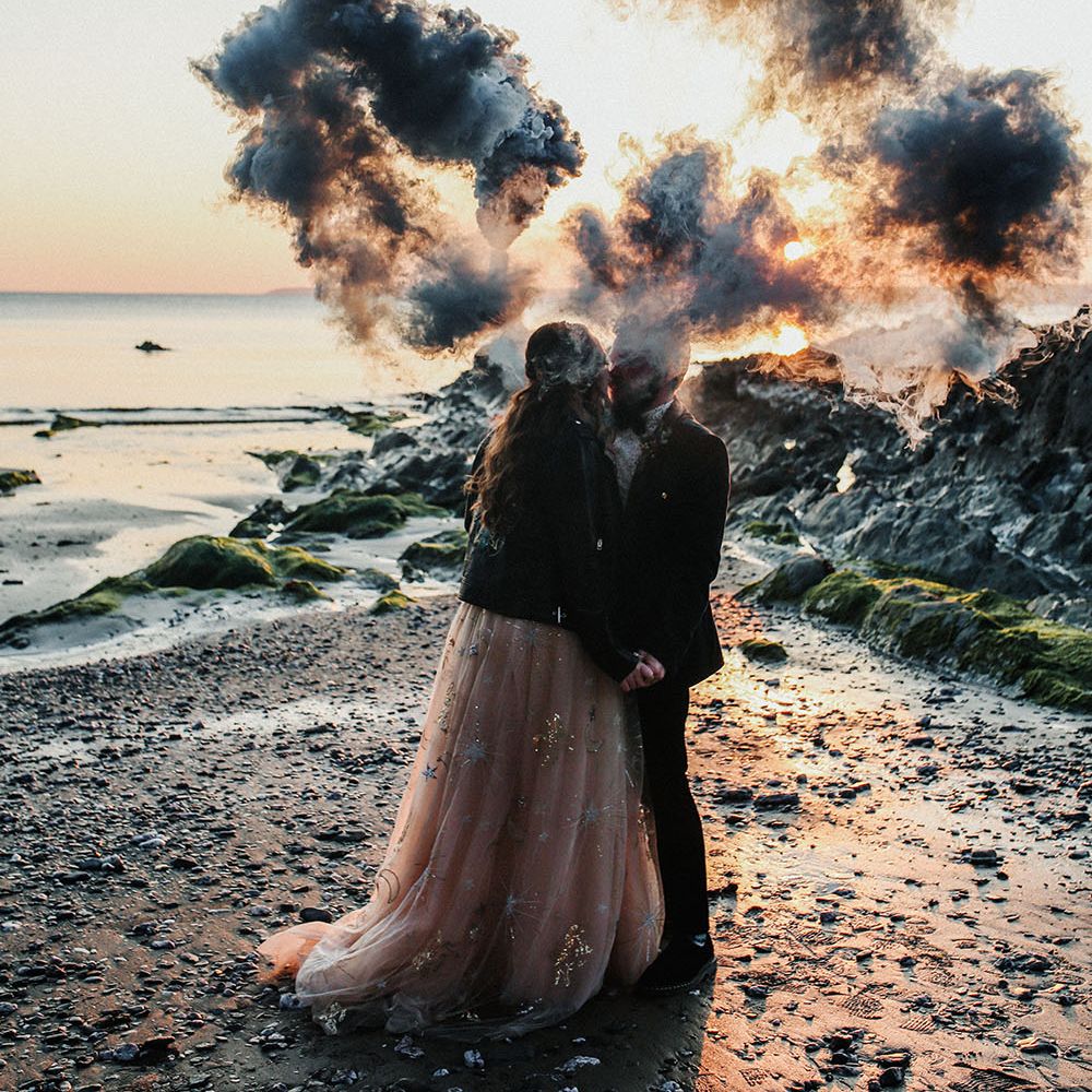 Bride & groom hold colourful smoke bombs on their wedding day along the beachfront in Cornwall 
