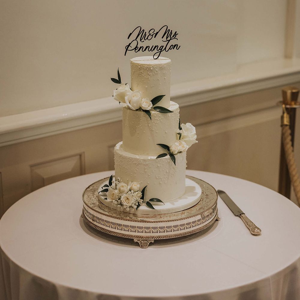 Three tier white frosted wedding cake with pearls and white rose decor and a personalised cake topper 
